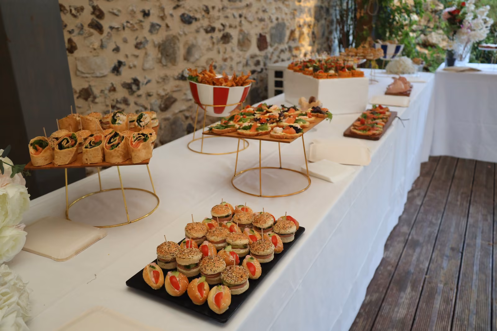 Buffet de cocktail dînatoire le long d'un mur en pierre : mini-burgers, wraps, toasts et présentoirs dorés