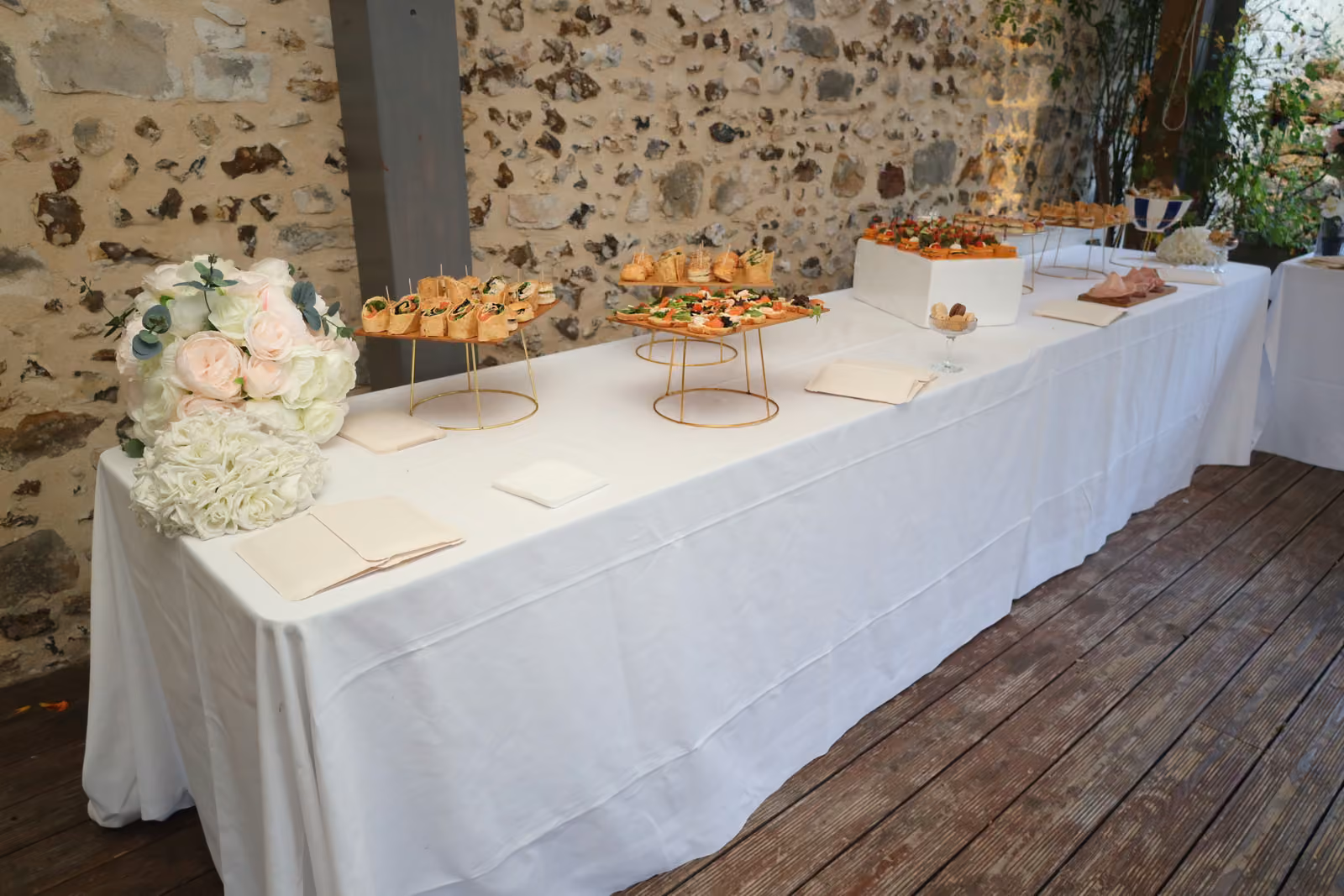 Buffet de cocktail sur nappe blanche en terrasse, présentoirs dorés garnis de bouchées et bouquet de roses