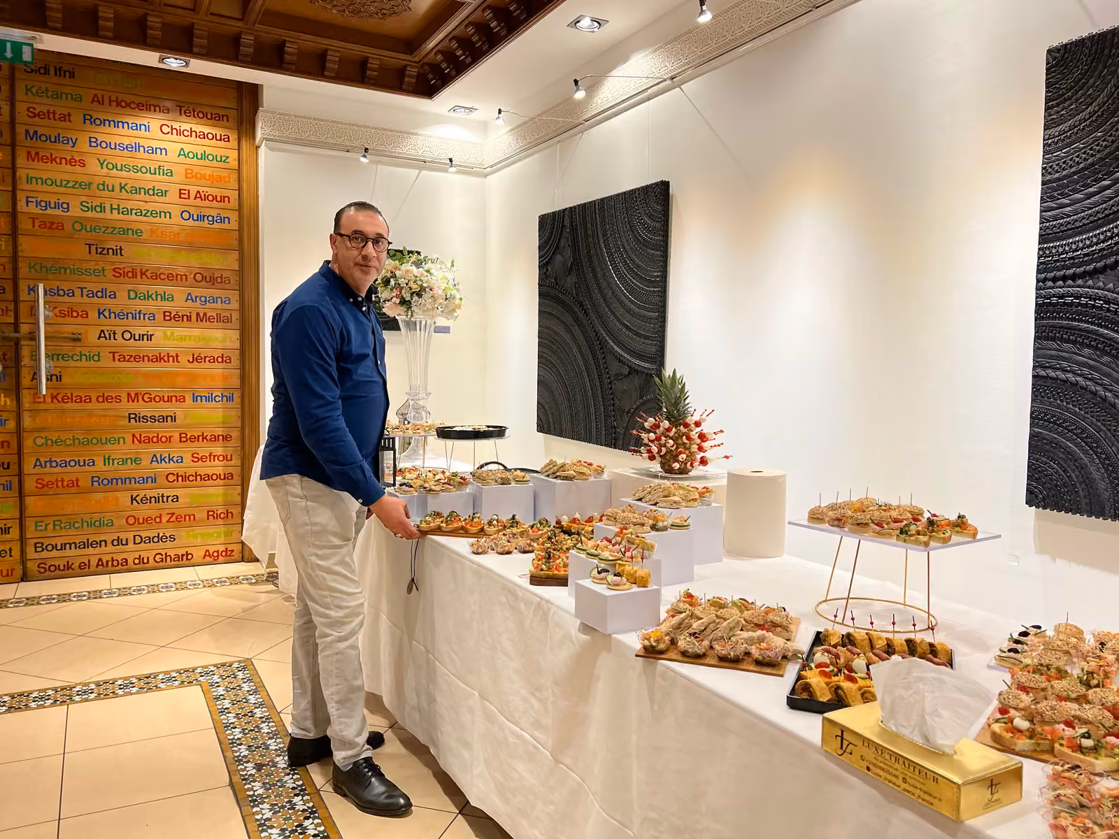 Chef traiteur en chemise bleue devant un buffet dînatoire dressé dans une salle de réception